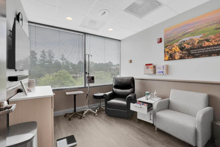 Modern clinic consultation room with a desk, chairs, and a small sofa seating area beside a large window with natural light.