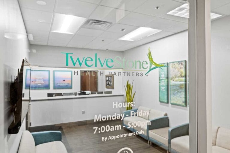 View through glass into TwelveStone Health Partners reception and waiting area, with the logo and office hours printed on the door.